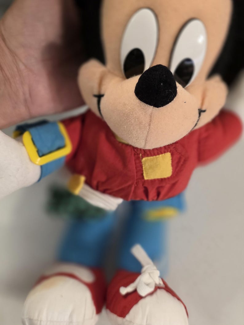 Peut inclure: Une peluche vintage de Mickey Mouse. La peluche porte une chemise rouge, un pantalon bleu et des chaussures rouges. Le jouet a un nez noir, de grands yeux noirs et une expression souriante. Le jouet est tenu par une personne.