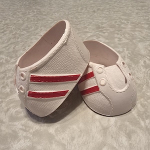 May include: A pair of white canvas sneakers with red stripes, designed for a doll. The shoes have a classic low-top design with a rounded toe and eyelets for laces. The red stripes add a pop of color to the otherwise plain white shoes.