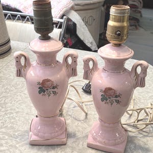 May include: Pair of vintage pink ceramic lamps with a floral rose design. Each lamp has a decorative handle on either side and a gold trim. The lamps have a square base and a brass lamp holder.