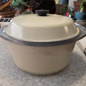 May include: Cream-colored vintage cooking pot with a lid and a black knob. The pot has a silver rim and handles. The pot is sitting on a light-colored surface. The pot is likely used for cooking.