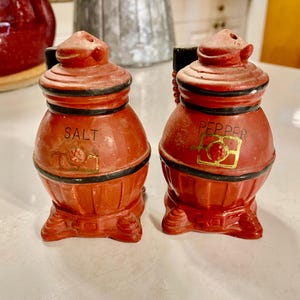 May include: A set of vintage red ceramic salt and pepper shakers shaped like a wood-burning stove. The shakers have black trim and gold lettering that reads "SALT" and "PEPPER".