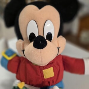 Peut inclure: Poup&eacute;e en peluche Mickey Mouse avec une veste rouge, des manches bleues et des oreilles noires. La poup&eacute;e a de grands yeux blancs et un nez noir. Un patch carr&eacute; jaune est sur la veste.