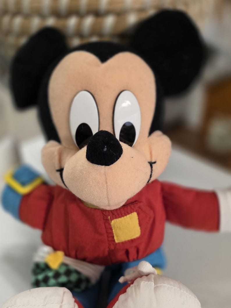 Peut inclure: Poup&eacute;e en peluche Mickey Mouse avec une veste rouge, des manches bleues et des oreilles noires. La poup&eacute;e a de grands yeux blancs et un nez noir. Un patch carr&eacute; jaune est sur la veste.