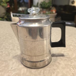 May include: A vintage aluminum coffee percolator with a clear glass knob on the lid and a black handle. The percolator has a silver finish and a spout for pouring. A classic kitchen appliance.