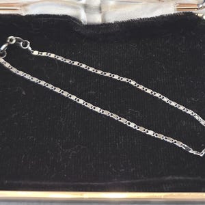 May include: A delicate silver chain bracelet with a repeating rectangular link design. The bracelet is displayed on a black velvet surface inside a small, open jewelry box. The box has a silver-colored metal frame.