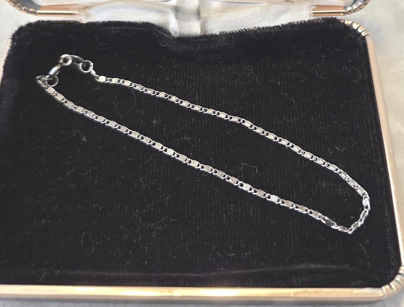 May include: A delicate silver chain bracelet with a repeating rectangular link design. The bracelet is displayed on a black velvet surface inside a small, open jewelry box. The box has a silver-colored metal frame.