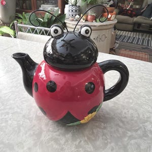 May include: A whimsical ladybug-shaped teapot. The teapot features a glossy black head with large white-eyed details and a red body with black spots. The handle and spout are black, and a small sunflower is painted on the bottom.