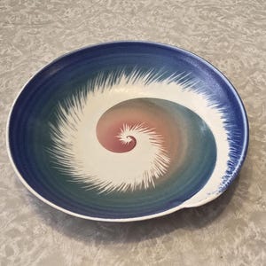 May include: A round ceramic bowl featuring a central spiral design that transitions from soft pink to muted green. The outer rim is a deep blue, with white, feathery, brush-like strokes radiating outward from the center toward the edge of the bowl.