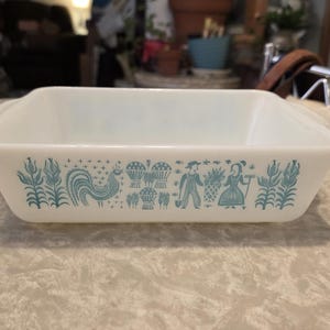 May include: Rectangular white Pyrex baking dish with a turquoise design featuring a rooster, people, and plants. The dish has two handles and is sitting on a patterned surface.