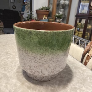 May include: A large, ceramic plant pot with a speckled white, green, and brown exterior. The pot has a wide opening and a slightly flared base. The interior is a solid brown color. The pot is sitting on a white surface.