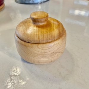 May include: A small, round wooden container with a lid. The container and lid are made of light-colored wood with a natural finish. The lid has a small, rounded knob for easy handling. The container is likely used for storing small items.