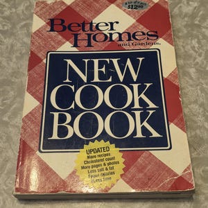 May include: A vintage cookbook titled "Better Homes and Gardens New Cook Book." The cover has a red and white checked pattern with a blue rectangle containing the title. A yellow sticker highlights updated features.