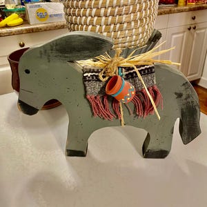 Peut inclure: Une figurine de donkey en bois gris avec une selle et un petit pot orange sur le dos. La selle est décorée d'une couverture tissée colorée et de glands rouges.