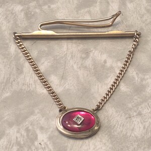 May include: A gold-toned tie clip with a chain and a decorative oval pendant. The pendant features a vibrant pink center with a small, square, clear stone. The clip is designed to secure a tie.