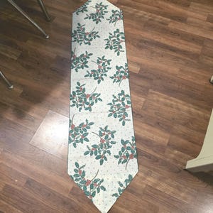 May include: A rectangular table runner with a cream background and a repeating pattern of green holly leaves and red berries. The runner has a gold tassel at each end. A festive holiday decoration.