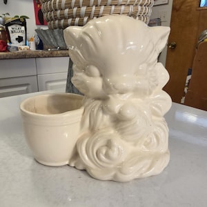 May include: Cream-colored ceramic cat planter with a small attached pot. The cat is in a seated position with its paws together. The planter has a glossy finish and is likely vintage.