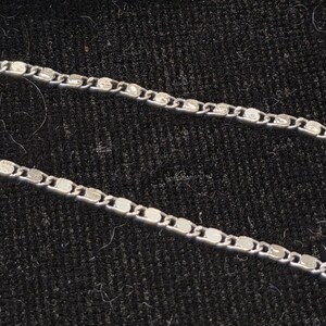 May include: Close-up of a silver necklace with a delicate, flat chain design. The chain is composed of small, interconnected, rectangular links. The necklace is displayed against a dark, textured background.