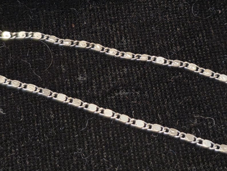 May include: Close-up of a silver necklace with a delicate, flat chain design. The chain is composed of small, interconnected, rectangular links. The necklace is displayed against a dark, textured background.