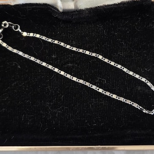 May include: A delicate silver chain bracelet with a repeating rectangular link design. The bracelet is displayed on a black velvet surface inside a gold-trimmed box. The clasp is visible at one end.