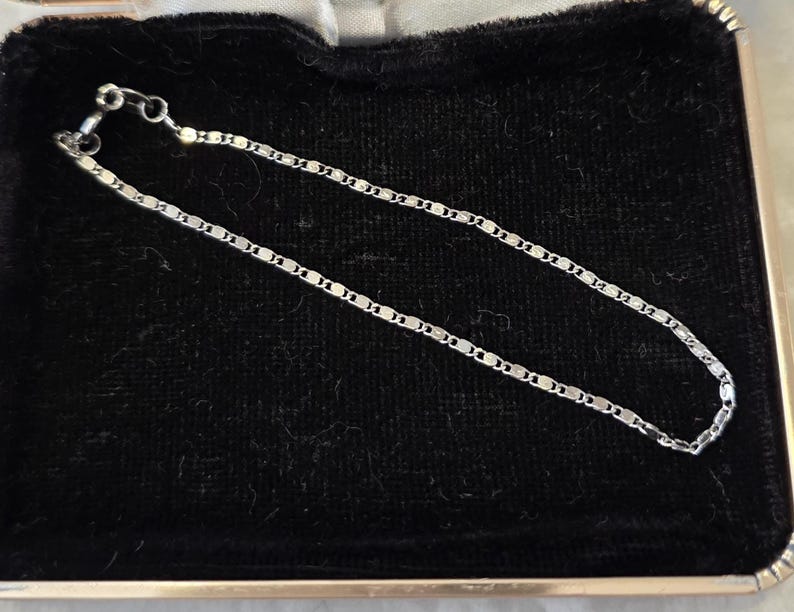 May include: A delicate silver chain bracelet with a repeating rectangular link design. The bracelet is displayed on a black velvet surface inside a gold-trimmed box. The clasp is visible at one end.