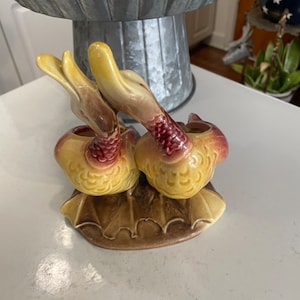 May include: A pair of yellow and brown ceramic duck planter figurines. The ducks are facing each other and have red accents on their wings. The ducks are sitting on a brown leaf-shaped base.