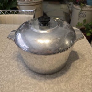 May include: A vintage aluminum cooking pot with a lid and a black knob. The pot has two side handles and is sitting on a light-colored surface. The pot is round and has a shiny, reflective surface.
