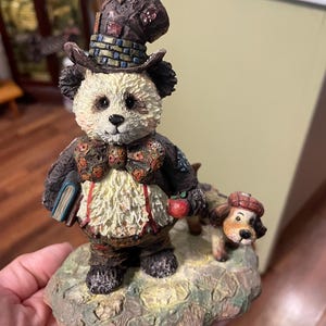 May include: A detailed figurine of a panda bear wearing a hat, bow tie, and suspenders, holding a book and an apple. A small dog with a hat stands beside the panda. The base is textured and painted with earth tones.