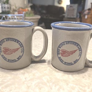 May include: Two off-white ceramic mugs with blue rims and handles. Each mug features a circular logo with a red wing design and the text "RED WING STONEWARE CO. RED WING, MN." The mugs are on a light-colored surface.