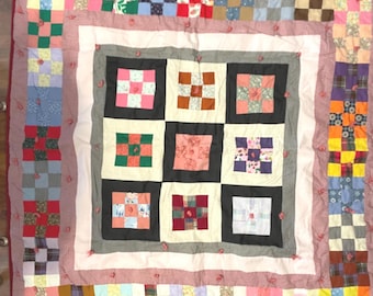 May include: A handmade patchwork quilt with a square pattern. The quilt features a variety of colorful fabric squares in shades of pink, blue, green, and yellow, bordered by a red and white dotted fabric. The quilt is a vintage item.