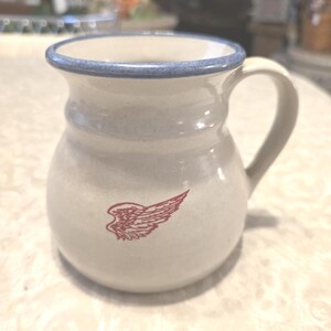May include: Cream-colored ceramic mug with a blue rim and handle. A red wing design is printed on the side. The mug has a rounded body and a slightly flared top.