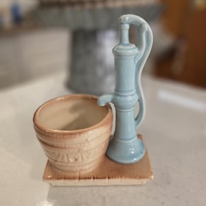 May include: A ceramic water pump and basin set. The pump is light blue, and the basin is beige with a decorative pattern. The set is mounted on a brown rectangular base. The background is blurred.