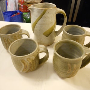 May include: Set of five pottery mugs and a pitcher with a brown, green, and white glaze. The mugs and pitcher have a rustic, handmade look.