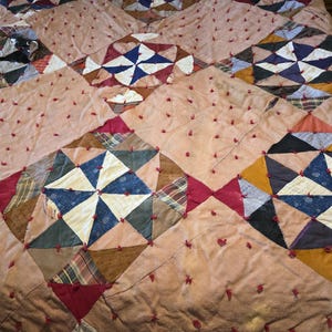 May include: A handmade quilt with a patchwork design. The quilt features geometric shapes in various colors, including blue, red, brown, and cream, arranged in a diamond pattern. The quilt is made of different fabrics and textures, with red knots throughout.