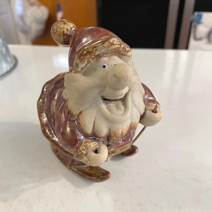 Puede incluir: Figura de cerámica de Papá Noel con una cara sonriente, vistiendo un atuendo y sombrero rojo y marrón. La figura tiene una cara y barba de color beige claro, y está diseñada para parecer que está sentado en esquís. Una decoración festiva.
