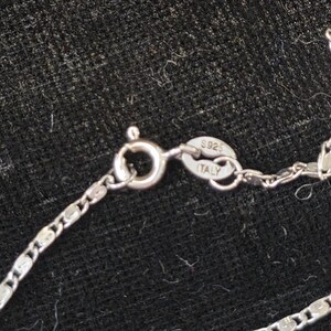 May include: Close-up of a silver necklace with a delicate chain and a lobster clasp. The clasp is connected to a chain with a series of small, flat, rectangular links. The clasp is stamped with "S925 ITALY".