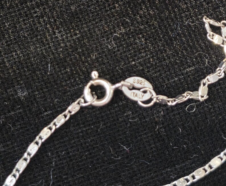 May include: Close-up of a silver necklace with a delicate chain and a lobster clasp. The clasp is connected to a chain with a series of small, flat, rectangular links. The clasp is stamped with "S925 ITALY".