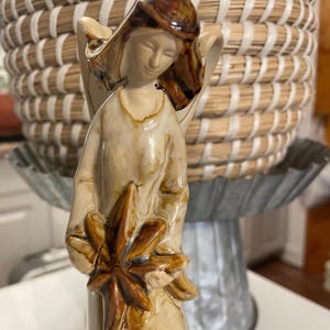 May include: A ceramic angel figurine with a serene expression, wearing a cream-coloured robe and holding a star-shaped object. The angel's hair is a dark brown, and the wings are visible. The figurine is set against a blurred background.