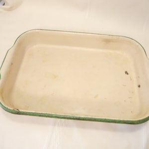 May include: Cream-coloured rectangular baking dish with a green rim. The dish has a slightly distressed appearance, suggesting it is vintage or well-used. It features small handles on the sides.