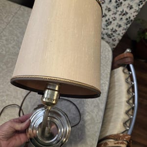 May include: A vintage table lamp with a beige fabric shade and a gold trim. The lamp has a brass-colored base and a brown cord. The lamp is designed to be placed on a table or desk.
