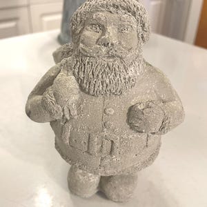 May include: A concrete Santa Claus figurine with a textured finish. The statue features a detailed beard, hat, and coat, and is holding a sack. The figure is in a standing position and is a festive holiday decoration.