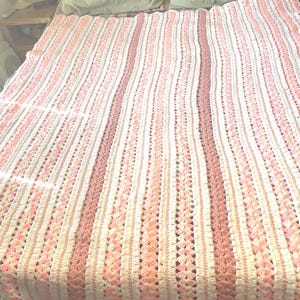May include: A handmade crochet blanket in shades of pink and white, with vertical stripes. The blanket is displayed on a bed with light green pillows. The crochet pattern features a textured design, adding visual interest to the soft, cozy blanket.