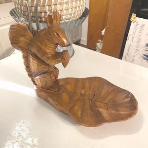 May include: A carved wooden squirrel figurine holding a nut, perched on a leaf-shaped dish. The squirrel is detailed with textured fur and a bushy tail. The dish is a warm brown color, perfect for holding small items.