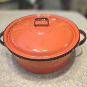 May include: An orange enamel pot with a matching lid. The pot has a black rim and handles, and a black handle on the lid. The pot is round and appears to be vintage.