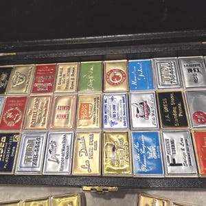 May include: A collection of vintage metal advertising signs, each with unique designs and company names. The signs are arranged in a black display case, showcasing a variety of colors and textures. Some signs feature embossed details.