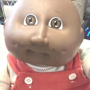 May include: A Cabbage Patch Kids doll with a smooth, round, dark-toned vinyl head, brown eyes, and dimpled cheeks. The doll wears a white shirt under red corduroy overalls featuring two white buttons on the front bib and a small green tag.