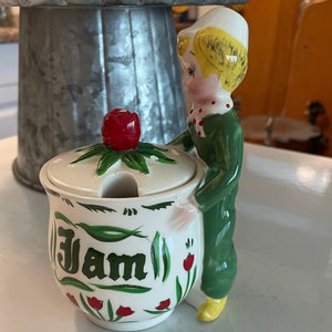 May include: A white ceramic jam jar with a green and white floral design and the word "Jam" written in green. A small, white-haired figurine with a green dress is holding the jar.