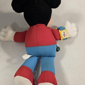 Peut inclure: Une peluche Mickey Mouse, vue de dos, portant une montre jaune avec un bracelet bleu. Le jouet a une chemise rouge, un pantalon bleu et des chaussures rouges. Les oreilles sont noires et les mains blanches.
