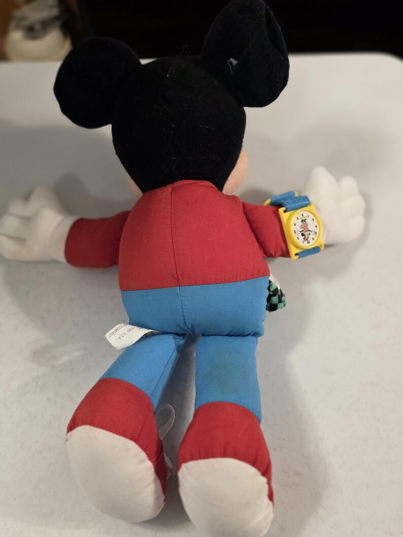 Peut inclure: Une peluche Mickey Mouse, vue de dos, portant une montre jaune avec un bracelet bleu. Le jouet a une chemise rouge, un pantalon bleu et des chaussures rouges. Les oreilles sont noires et les mains blanches.