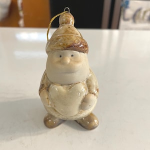 May include: A ceramic Christmas ornament shaped like a snowman. The snowman has a beige body, a brown hat, and a gold string for hanging. The ornament has a heart-shaped belly and small black eyes.