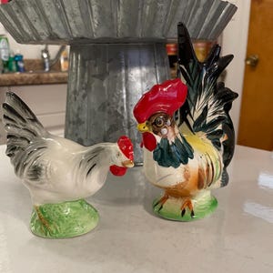 May include: Two ceramic rooster figurines. One rooster is white with black accents and the other is multi-colored with a red comb and black, green, and yellow accents.